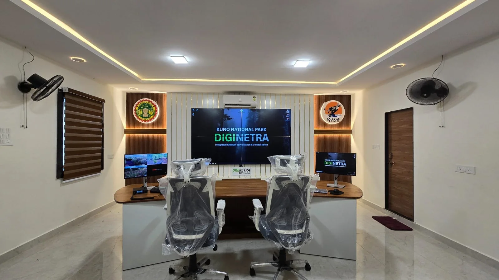 The DigiNetra control room inside Kuno National Park: two operator workstations facing a wall-mounted display reading “Kuno National Park DigiNetra — Integrated Cheetah Surveillance & Control Room”, with live camera feeds running on a side monitor.