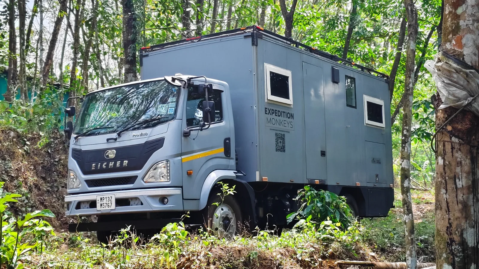 Bhanumati expedition camper truck parked among tall forest trees, front three-quarter view.